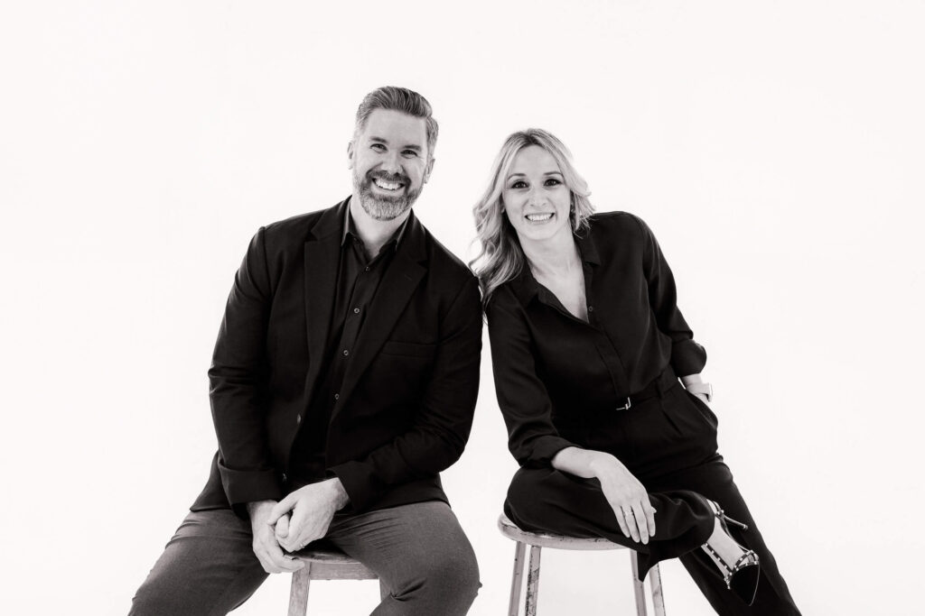 Black-and-white studio portrait of a couple seated on stools, photographed in a clean, editorial style with soft lighting and a relaxed, natural expression.