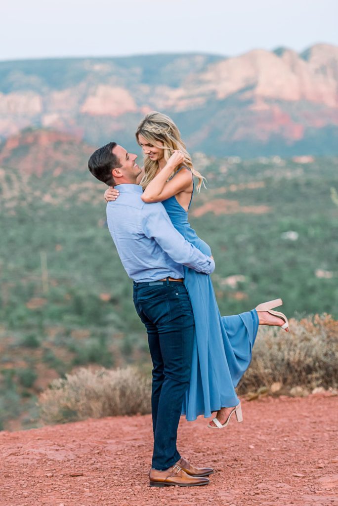He lifts her up looking at her along the edge of an overlook that shows Cathedral Rock in Sedona.