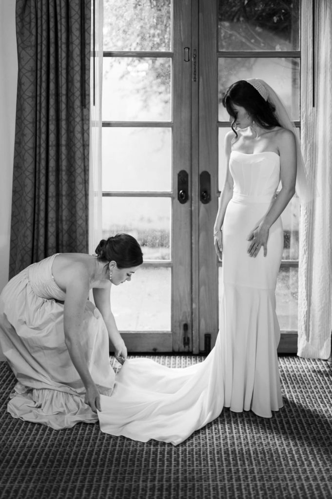 Bride standing in front of large window in wedding dress with bridesmaid fixing the train