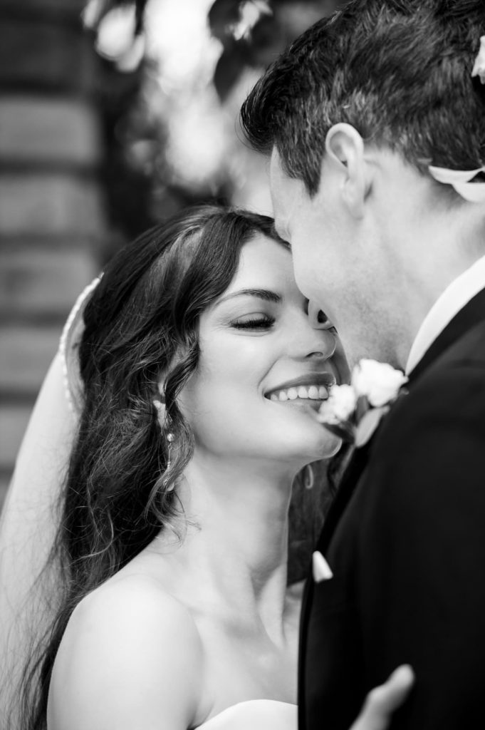 Bride smiling, while groom is about to kiss her in black and white