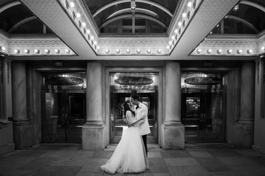 Bride and Groom kissing at Mayflower Hotel wedding venue at night in the main entrance on the sidewalk looking at the doors and building behind them
