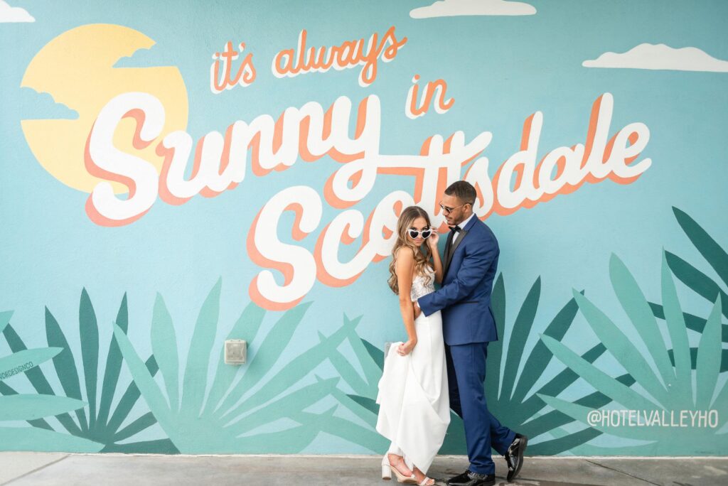 Bride and Groom standing in front of large mural that says "It's always sunny in Scottsdale" at Hotel Valley Ho.