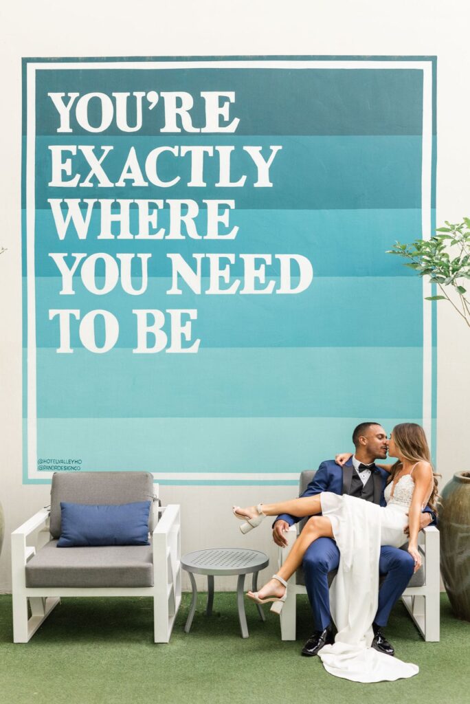 Bride sitting on the lap of the groom in a chair in front of a large mural that says "You're exactly where you need to be."