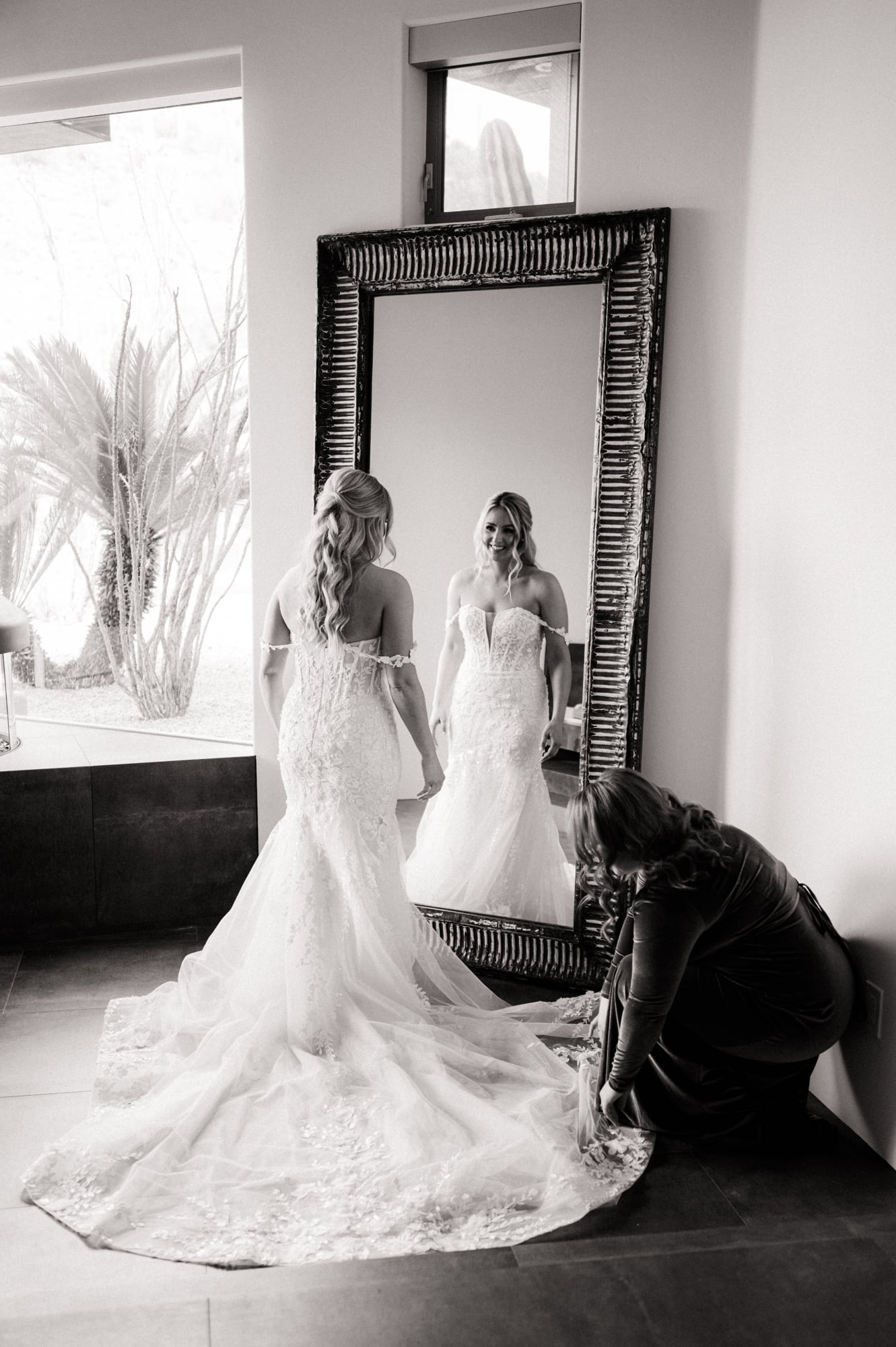 Bride in wedding dress looks in a large mirror in her getting ready room while a bridesmaid pulls out the train.